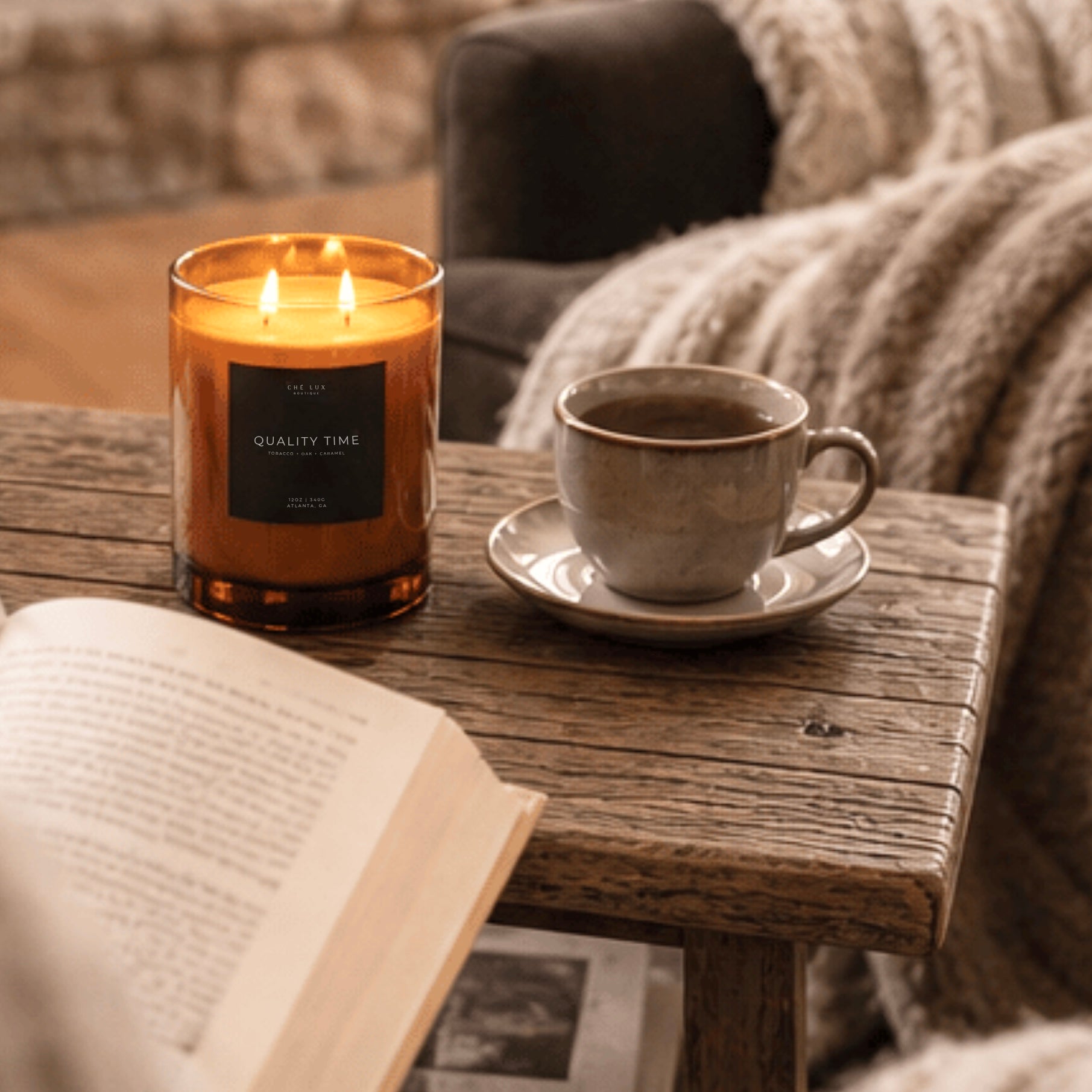 A cozy living room scene with a beige armchair draped in a chunky knit cream throw blanket, a round wooden coffee table with a book, mug, and lit candle, warm ambient lighting, and a window in the background.