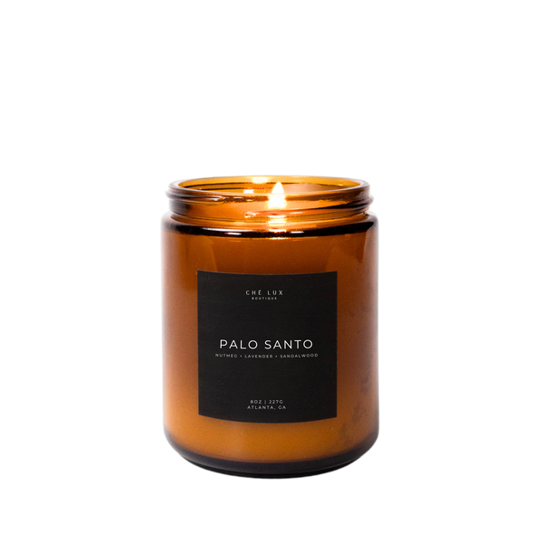 Che Lux Boutique product photo of a lit Palo Santo candle in an amber glass jar with a black label featuring nutmeg lavender sandalwood scents from Atlanta GA