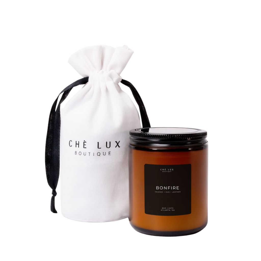 Product photography of a Che Lux Boutique Bonfire scented candle in an amber glass jar with a black lid and Incense Oud Leather notes, presented with a white drawstring bag featuring the brand logo and black ribbon from Atlanta GA