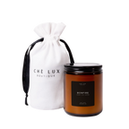 Product photography of a Che Lux Boutique Bonfire scented candle in an amber glass jar with a black lid and Incense Oud Leather notes, presented with a white drawstring bag featuring the brand logo and black ribbon from Atlanta GA