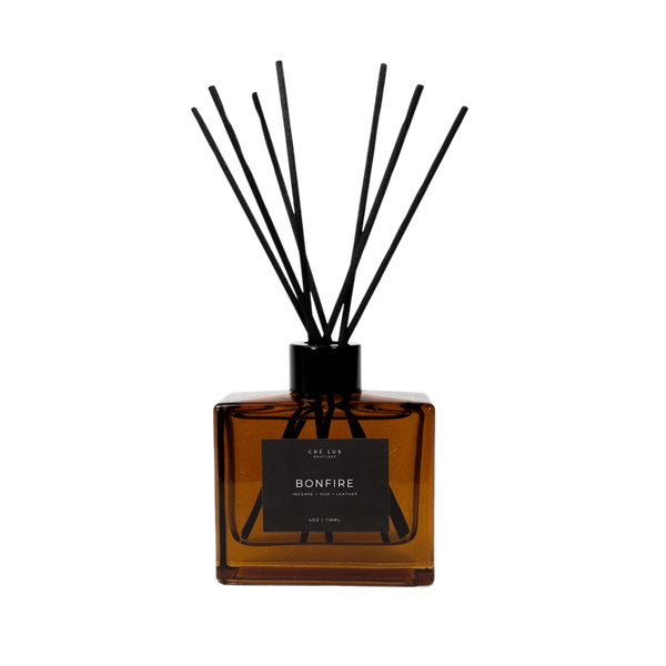 Ché Lux Boutique Bonfire reed diffuser with an amber glass bottle, black label detailing incense oud leather scent, and multiple black diffuser reeds on a black background