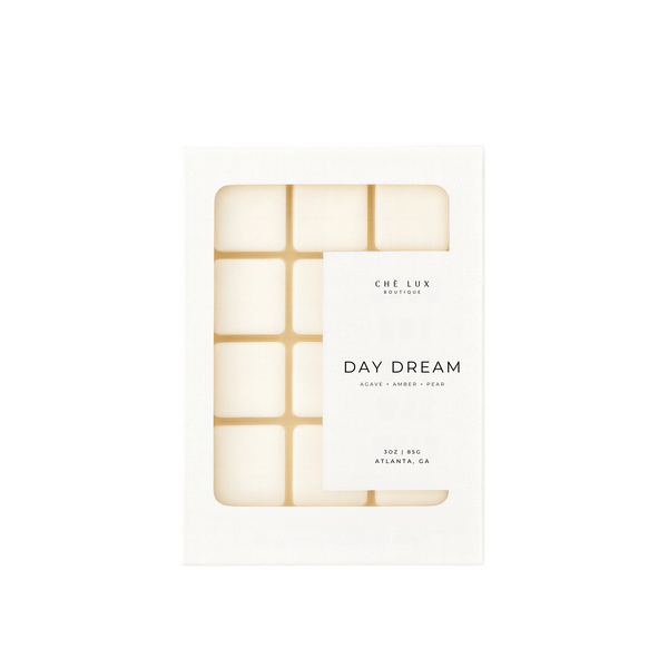 Product photography of CHÈ LUX BOUTIQUE "DAY DREAM" scented wax melts from Atlanta, GA in a white box with a clear window displaying twelve light-colored square melts featuring agave amber and pear fragrance notes