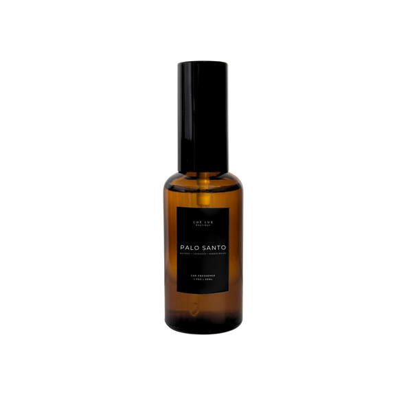 Product photograph by CHÉ LUX BOUTIQUE of an amber glass spray bottle of Palo Santo car freshener with a black label featuring white text listing nutmeg lavender and sandalwood notes and a black pump top