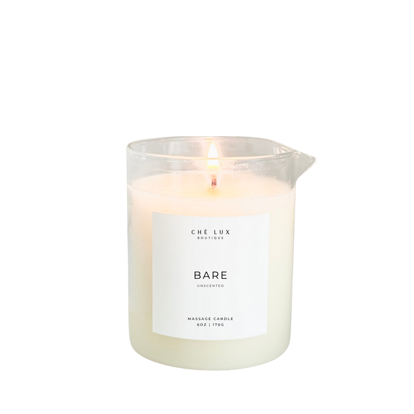 Digital photograph of Che Lux Boutique's Bare Unscented Massage Candle glowing with a warm flame in a clear glass container with a pouring spout, showcasing creamy off-white wax against a black background