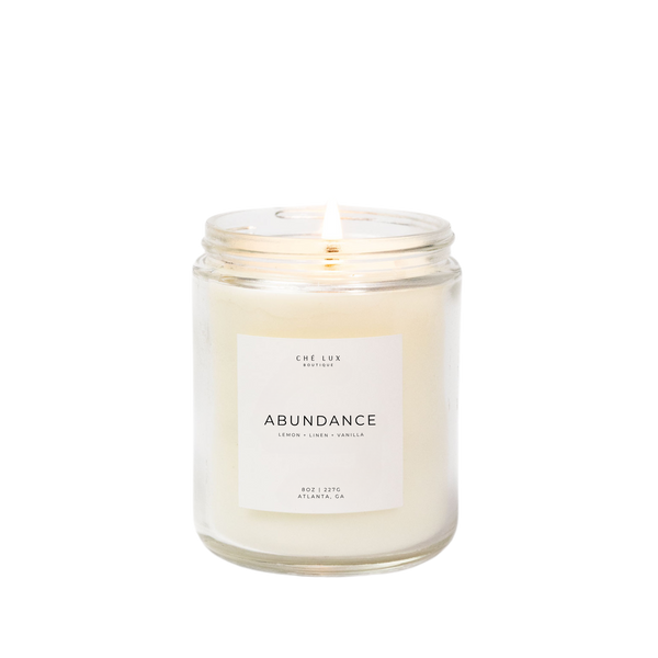 Photograph of a Ché Lux Boutique Abundance candle from Atlanta GA in a clear glass jar with a lit wick and a label detailing a lemon linen vanilla scent and 8oz 227g