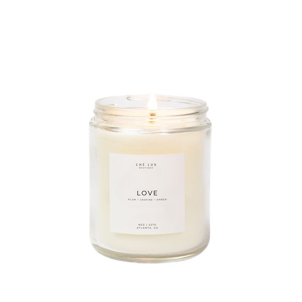 Photograph by Ché Lux Boutique of a lit white wax candle in a clear glass jar from Atlanta, GA featuring a prominent white label with "LOVE" and "PLUM + JASMINE + AMBER" scent details
