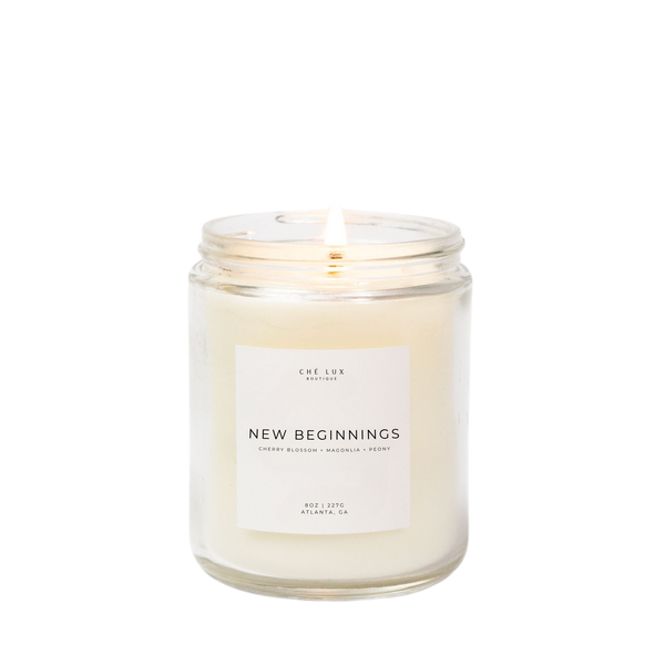Product photo by Ché Lux Boutique of a lit 'New Beginnings' candle in a clear glass jar containing creamy white wax with a label listing cherry blossom magnolia peony scents from Atlanta GA