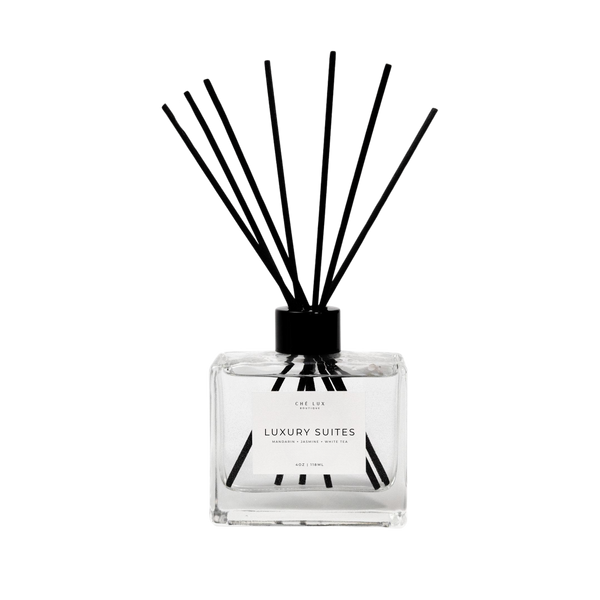 Product photo by CHÉ LUX BOUTIQUE of a clear square reed diffuser with black reeds and a white label indicating Luxury Suites scented Mandarin Jasmine White Tea on a black background