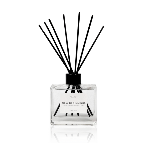Product photography by CHÉ LUX BOUTIQUE a minimalist square clear glass reed diffuser with black sticks and a white label featuring New Beginnings cherry blossom magnolia peony scent on a reflective black background