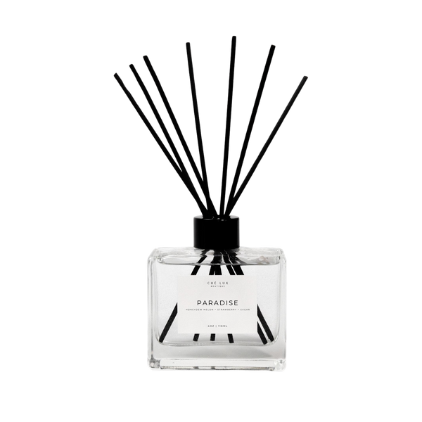 Ché Lux Boutique product photo of a square clear glass Paradise reed diffuser featuring black reeds and a white label describing a Honeydew Melon Strawberry Sugar scent