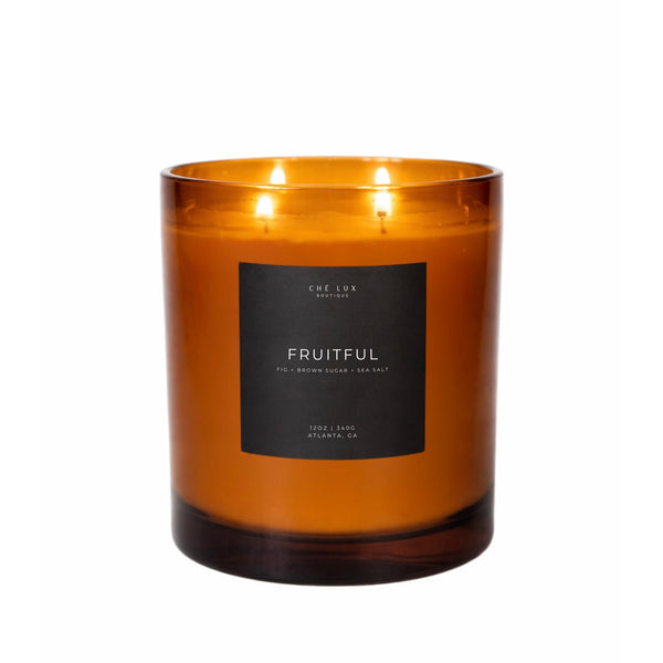 Product photo: Ché Lux Boutique's FRUITFUL three-wick candle in an amber glass jar with three lit wicks, featuring a black label detailing fig, brown sugar, and sea salt scent notes from Atlanta GA