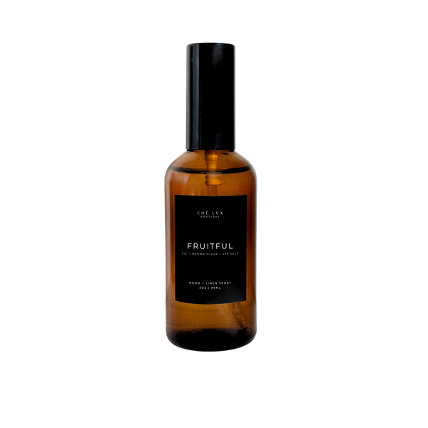 Product photo by Ché Lux Boutique of an amber glass bottle of Fruitful room and linen spray with a black label detailing fig brown sugar sea salt scent notes and a black spray top