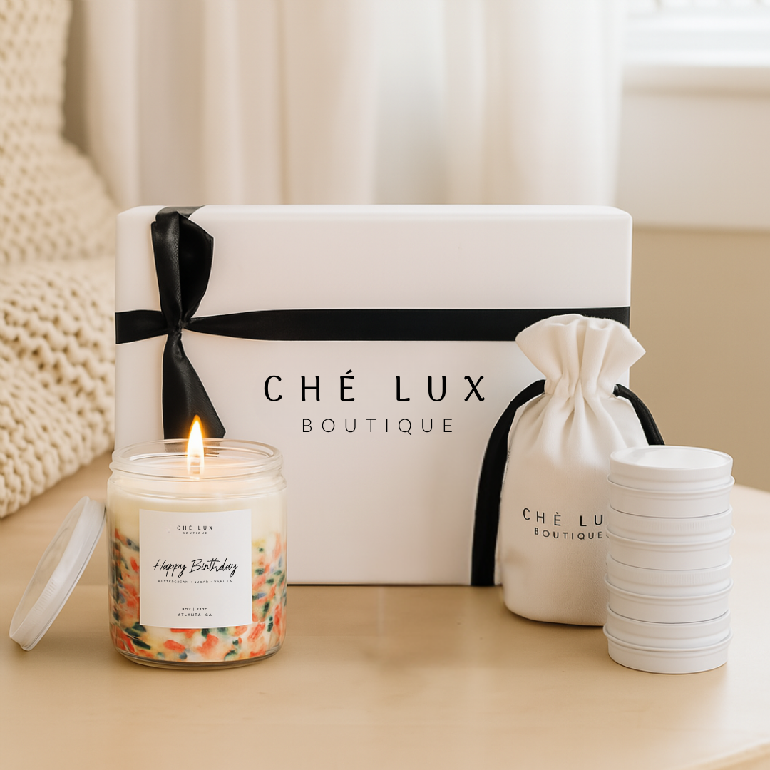 Candle with a 'Happy Birthday' label, 'Che Lux Boutique' box, and drawstring bag on a neutral background.