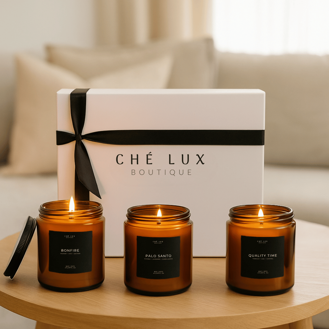 Three lit candles with labels in front of a 'Ché Lux Boutique' box on a table.